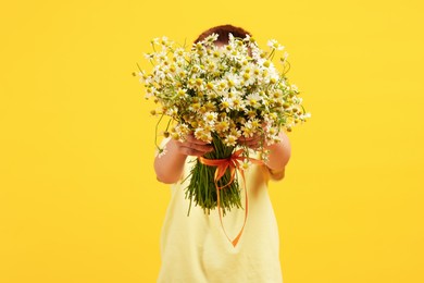Photo of Little boy hiding behind bouquet of beautiful chamomile flowers on yellow background