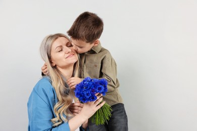 Photo of Little boy kissing his smiling mother with bouquet of beautiful cornflowers on light grey background. Space for text