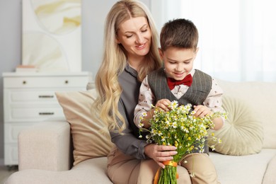 Photo of Little boy and his smiling mother with bouquet of beautiful chamomile flowers on sofa at home
