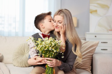 Photo of Little boy kissing his mother with bouquet of beautiful chamomile flowers on sofa at home