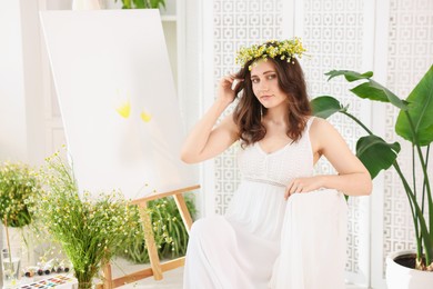 Photo of Beautiful woman with chamomile wreath near easel indoors