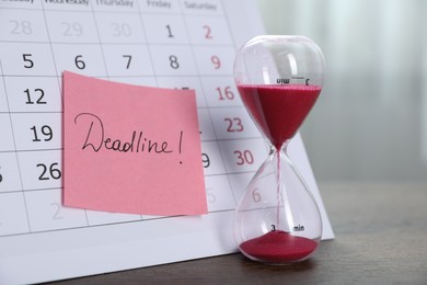 Photo of Sticky note with word Deadline, calendar and hourglass with flowing sand on wooden table indoors, closeup