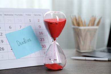 Photo of Sticky note with word Deadline, calendar and hourglass with flowing sand on table indoors, closeup
