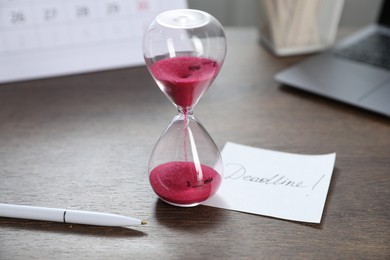 Photo of Sticky note with word Deadline, hourglass and pen on wooden table, closeup