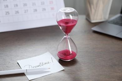 Photo of Sticky note with word Deadline, hourglass and pen on wooden table, closeup