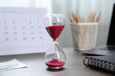 Photo of Deadline. Hourglass with flowing sand, stationery, calendar and laptop on wooden table indoors