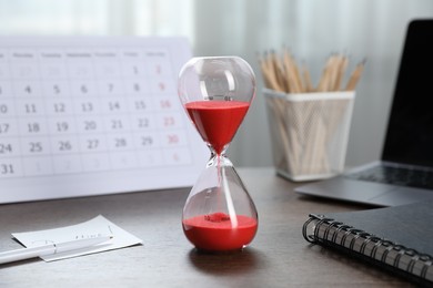 Photo of Deadline. Hourglass with flowing sand, stationery, calendar and laptop on wooden table indoors