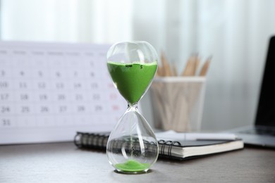 Photo of Deadline. Hourglass with flowing sand, stationery, calendar and laptop on wooden table indoors