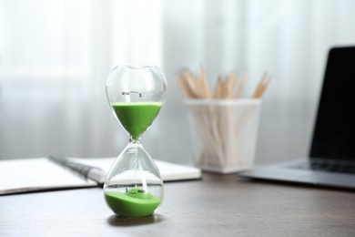 Photo of Deadline. Hourglass with flowing sand, stationery and laptop on wooden table indoors, space for text