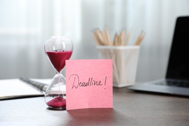 Photo of Sticky note with word Deadline, hourglass, stationery and laptop on wooden table indoors