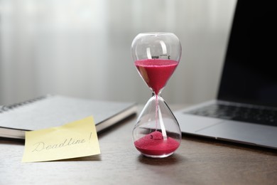 Photo of Sticky note with word Deadline, hourglass, notebook and laptop on wooden table indoors, closeup