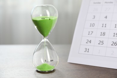 Photo of Deadline. Hourglass with flowing sand and calendar on wooden table indoors, closeup