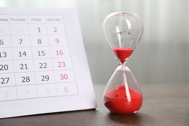 Photo of Deadline. Hourglass with flowing sand and calendar on wooden table indoors, closeup