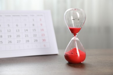 Photo of Deadline. Hourglass with flowing sand and calendar on wooden table indoors