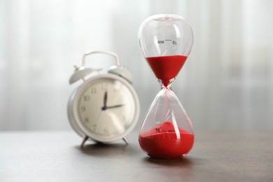 Photo of Deadline. Hourglass with flowing sand and alarm clock on wooden table indoors