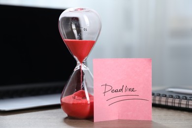 Photo of Sticky note with word Deadline, hourglass, notebook and laptop on wooden table indoors, closeup