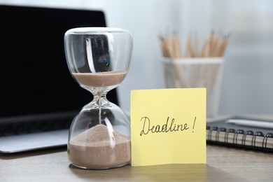 Photo of Sticky note with word Deadline, hourglass, stationery and laptop on wooden table indoors, closeup