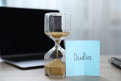 Photo of Sticky note with word Deadline, hourglass and laptop on wooden table indoors, closeup