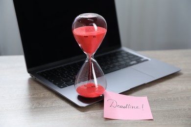 Photo of Sticky note with word Deadline, hourglass and laptop on wooden table indoors, closeup