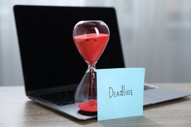 Photo of Sticky note with word Deadline, hourglass and laptop on wooden table indoors, closeup