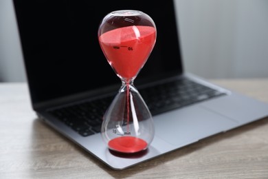 Photo of Deadline. Hourglass with flowing sand and laptop on wooden table indoors, closeup