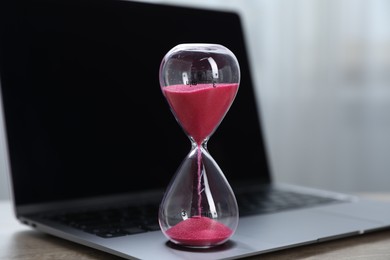 Photo of Deadline. Hourglass with flowing sand and laptop on table indoors, closeup