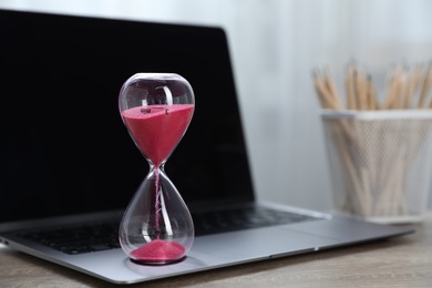 Photo of Deadline. Hourglass with flowing sand, stationery and laptop on wooden table indoors, closeup