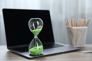 Photo of Deadline. Hourglass with flowing sand, stationery and laptop on wooden table indoors