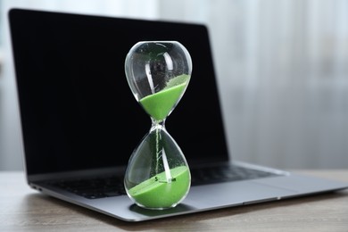 Photo of Deadline. Hourglass with flowing sand and laptop on wooden table indoors, closeup