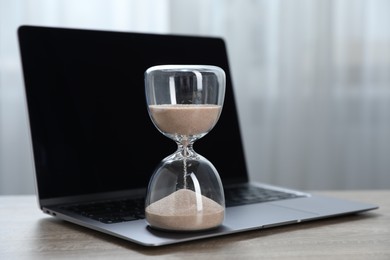 Photo of Deadline. Hourglass with flowing sand and laptop on wooden table indoors, closeup