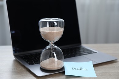 Photo of Sticky note with word Deadline, hourglass and laptop on wooden table indoors, closeup