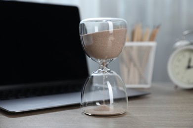 Photo of Deadline. Hourglass with flowing sand, stationery, laptop and alarm clock on wooden table indoors, closeup