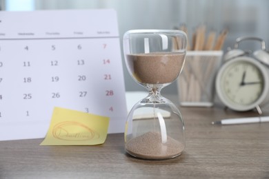 Photo of Sticky note with word Deadline, hourglass, calendar, stationery and alarm clock on wooden table indoors