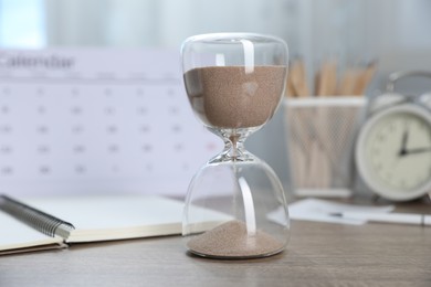 Photo of Deadline. Hourglass with flowing sand, stationery, calendar and alarm clock on wooden table indoors