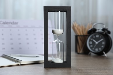 Photo of Deadline. Hourglass with flowing sand, stationery, calendar and alarm clock on wooden table indoors