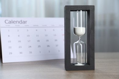 Photo of Deadline. Hourglass with flowing sand and calendar on wooden table indoors