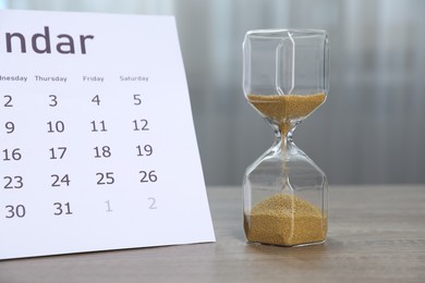 Photo of Deadline. Hourglass with flowing sand and calendar on wooden table indoors, closeup