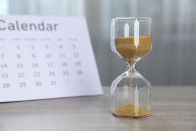 Photo of Deadline. Hourglass with flowing sand and calendar on wooden table indoors