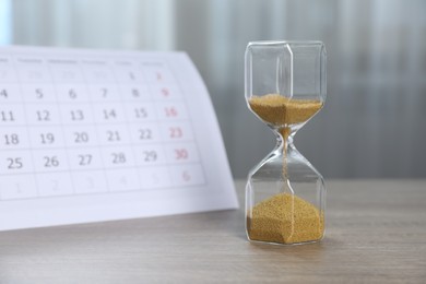 Photo of Deadline. Hourglass with flowing sand and calendar on wooden table indoors