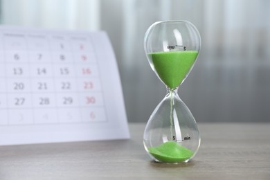 Photo of Deadline. Hourglass with flowing sand and calendar on wooden table indoors