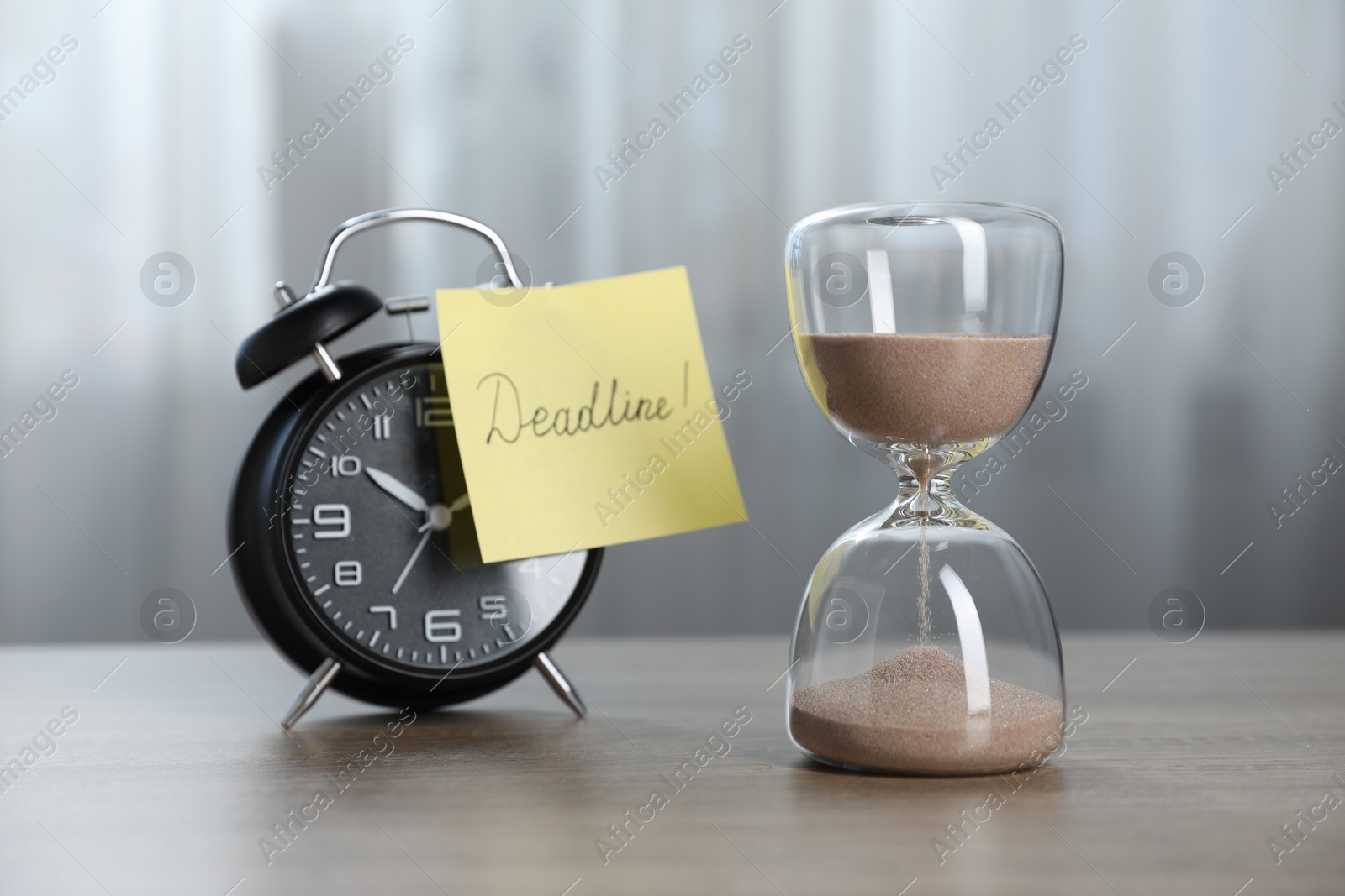 Sticky note with word Deadline, hourglass and alarm clock on wooden table indoors Photo of Sticky note with word Deadline, hourglass and alarm clock on wooden table indoors