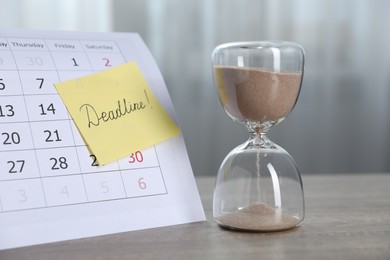 Photo of Sticky note with word Deadline, hourglass and calendar on wooden table indoors, closeup