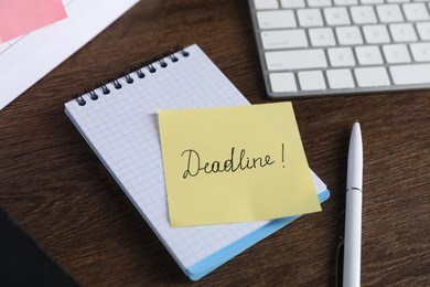Photo of Sticky note with word Deadline, notebook, pen and computer keyboard on wooden table, closeup