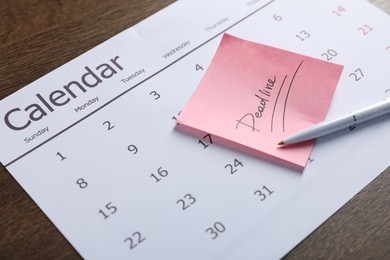 Photo of Sticky note with word Deadline, calendar and pen on wooden table, closeup