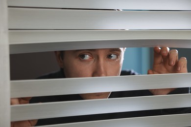 Photo of Paranoia. Worried man looking through window blinds indoors