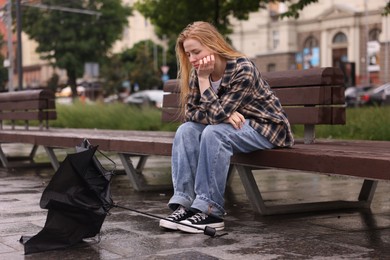 Photo of Frustrated woman sitting near broken umbrella on bench outdoors
