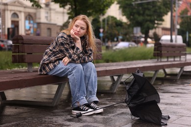 Photo of Frustrated woman sitting near broken umbrella on bench outdoors
