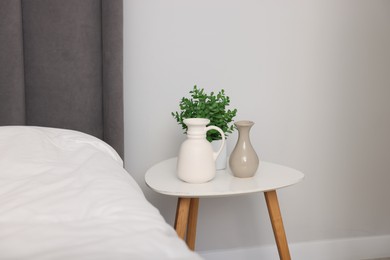 Photo of Beautiful vases and houseplant on white table near bed indoors
