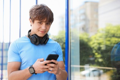 Photo of Cute teenage boy with headphones and smartphone near building outdoors