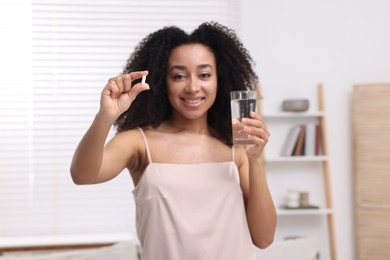 Photo of Beautiful Brazilian woman with glass of water and collagen pill indoors, selective focus